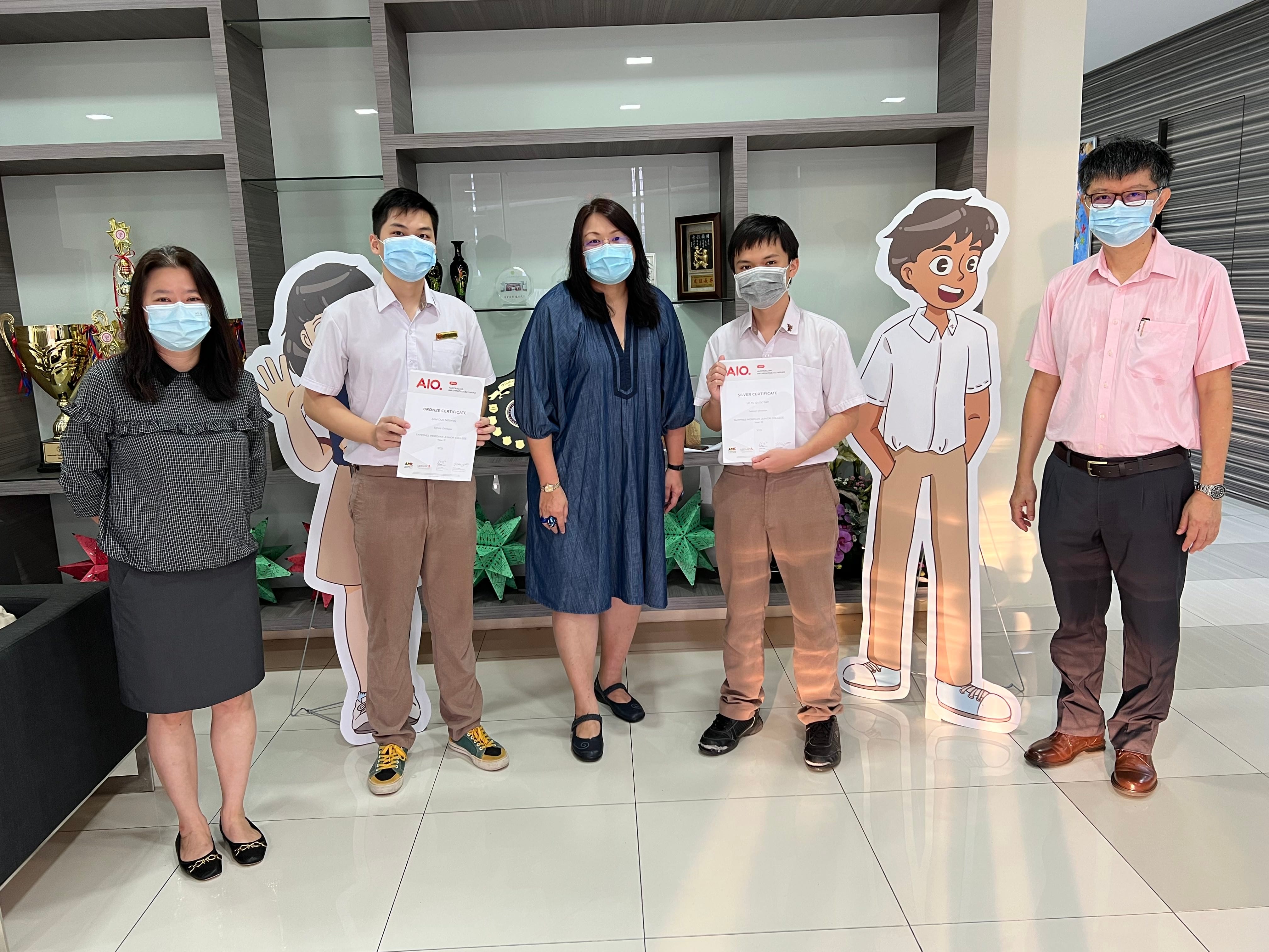 Five masked people pose indoors; two students hold AIO certificates, flanked by cartoon student cutouts and trophies nearby.